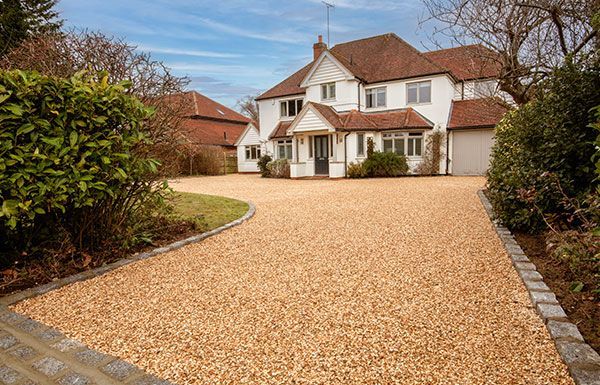 Gravel driveway leads to a white house with red roof tiles. Green bushes line the sides.