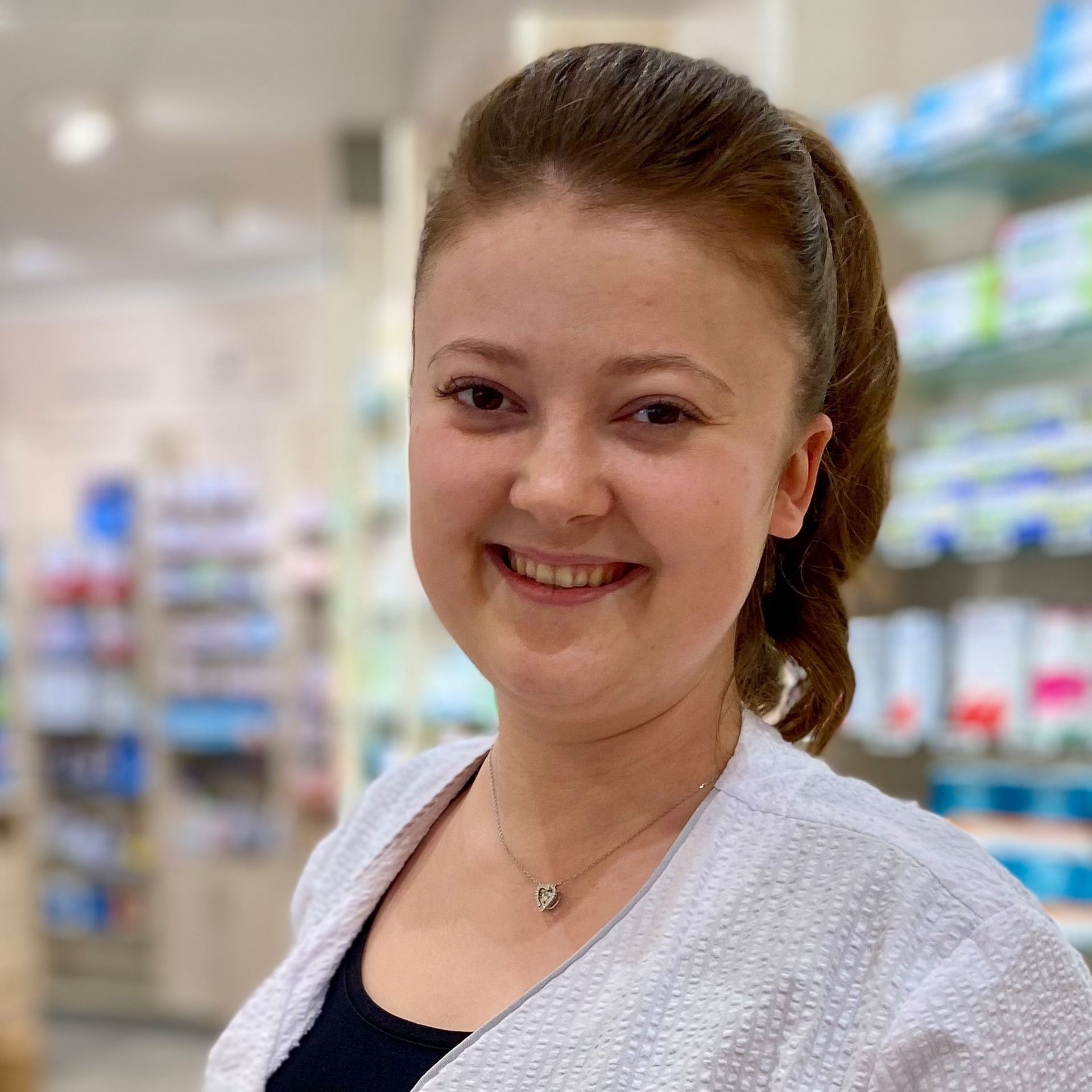 Eine Frau mit einer weißen Jacke und einer Halskette lächelt in einer Apotheke.