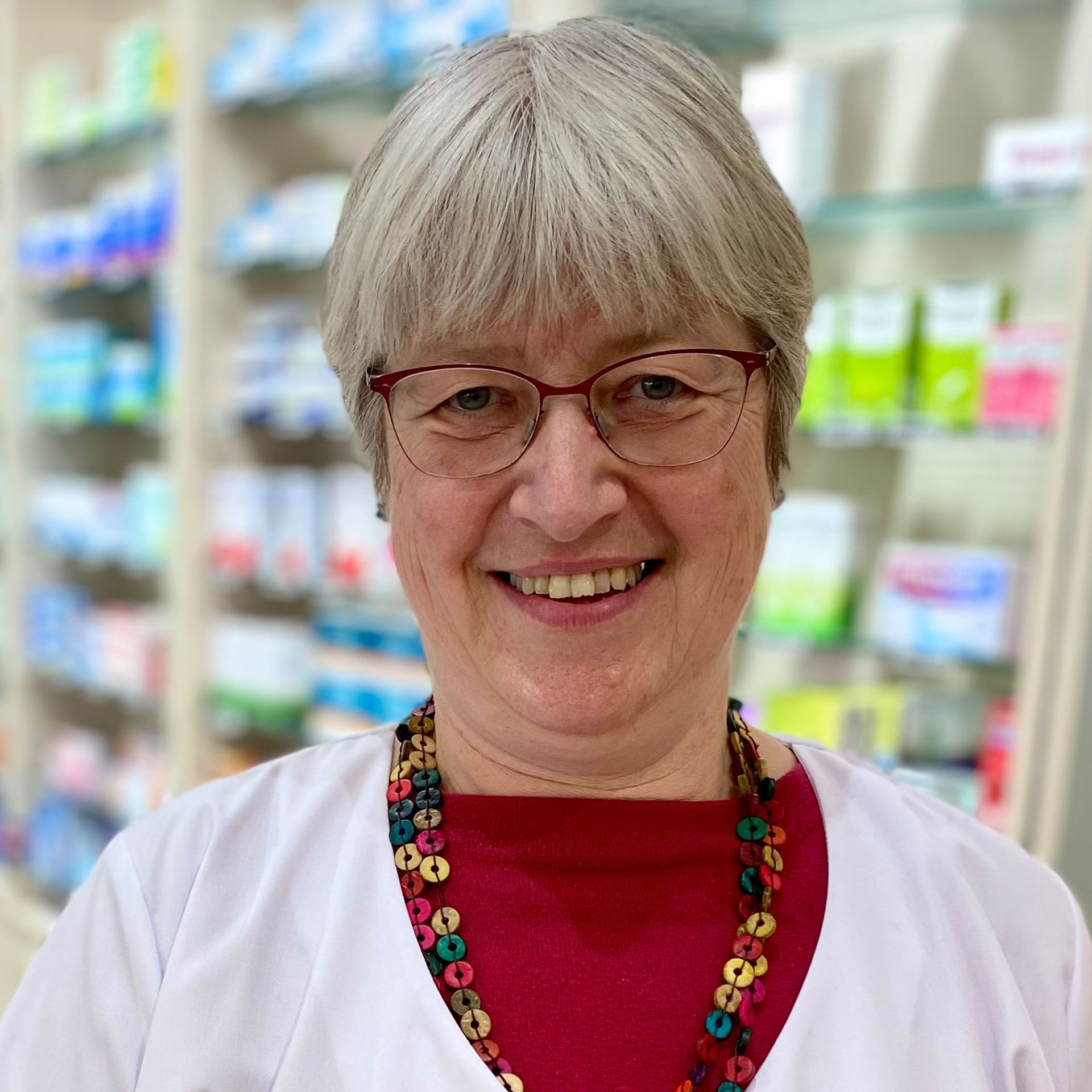 Eine Frau mit Brille und rotem Hemd lächelt in einer Apotheke.