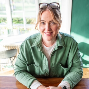 Woman with glasses atop head, smiling at a table, wearing a green jacket over a white shirt.