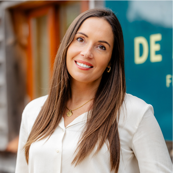 Woman with long brown hair, smiling, wearing a cream blouse, gold necklace; standing outdoors near a building.