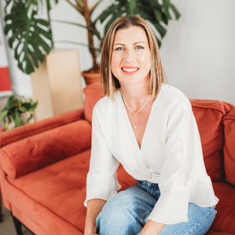 Woman smiles while sitting on an orange couch, wearing a white blouse and blue jeans. Lush plants in the background.
