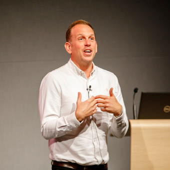 Man in white shirt speaking at a podium, gesturing with hands; laptop, microphone, gray background.