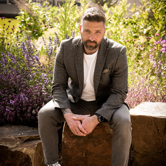 Man in gray blazer sits on a rock, hands clasped. He is in a garden with purple flowers.