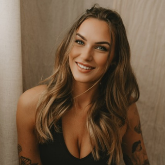 Woman with long wavy hair, smiling, wearing a black top, leaning against a wall.