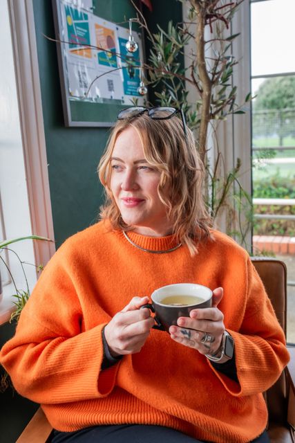 Woman in orange sweater holding a mug, looking out a window.