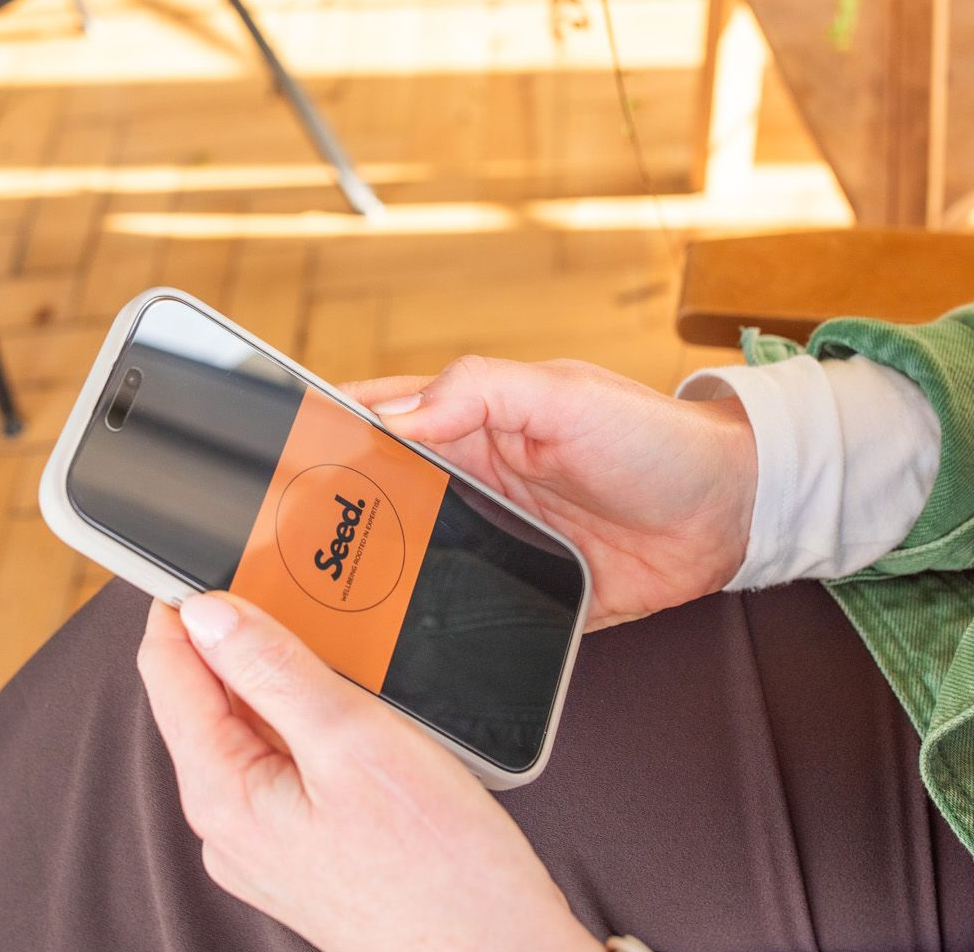 Person holding a smartphone with a logo on the screen, seated with brown pants.