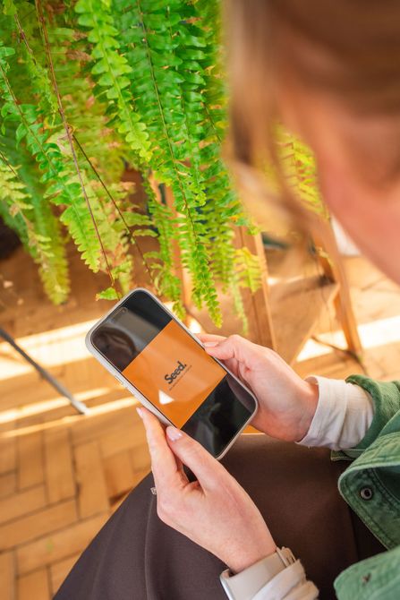 Person holding a phone with an app featuring a logo against a backdrop of green plants.