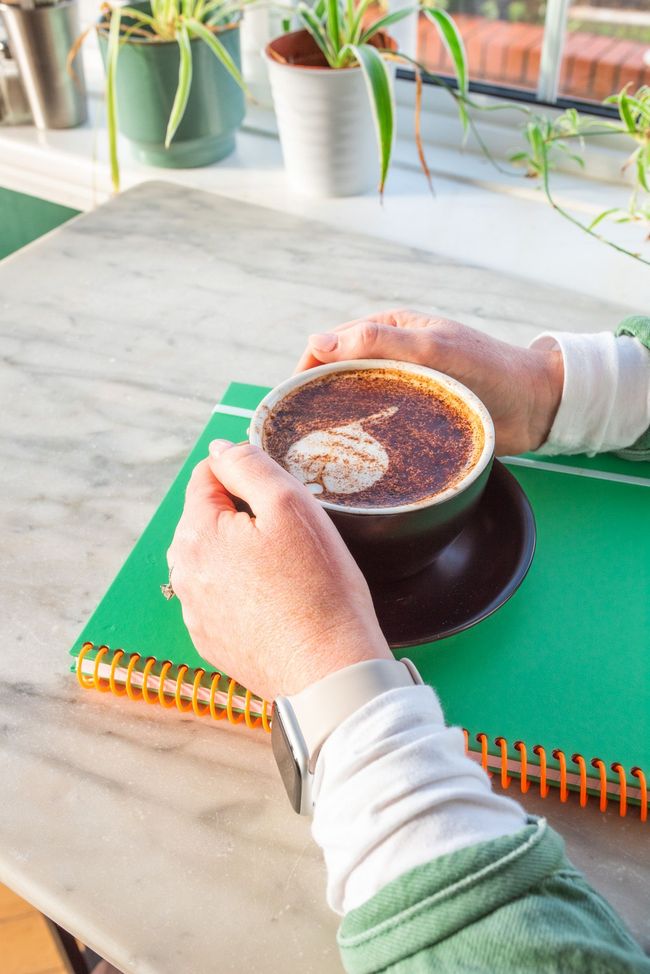 Hands holding a coffee cup on a green notebook, sunlight on a marble table with plants in the background.