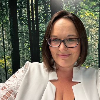 Woman in glasses and white blazer, smiling before a forest backdrop.
