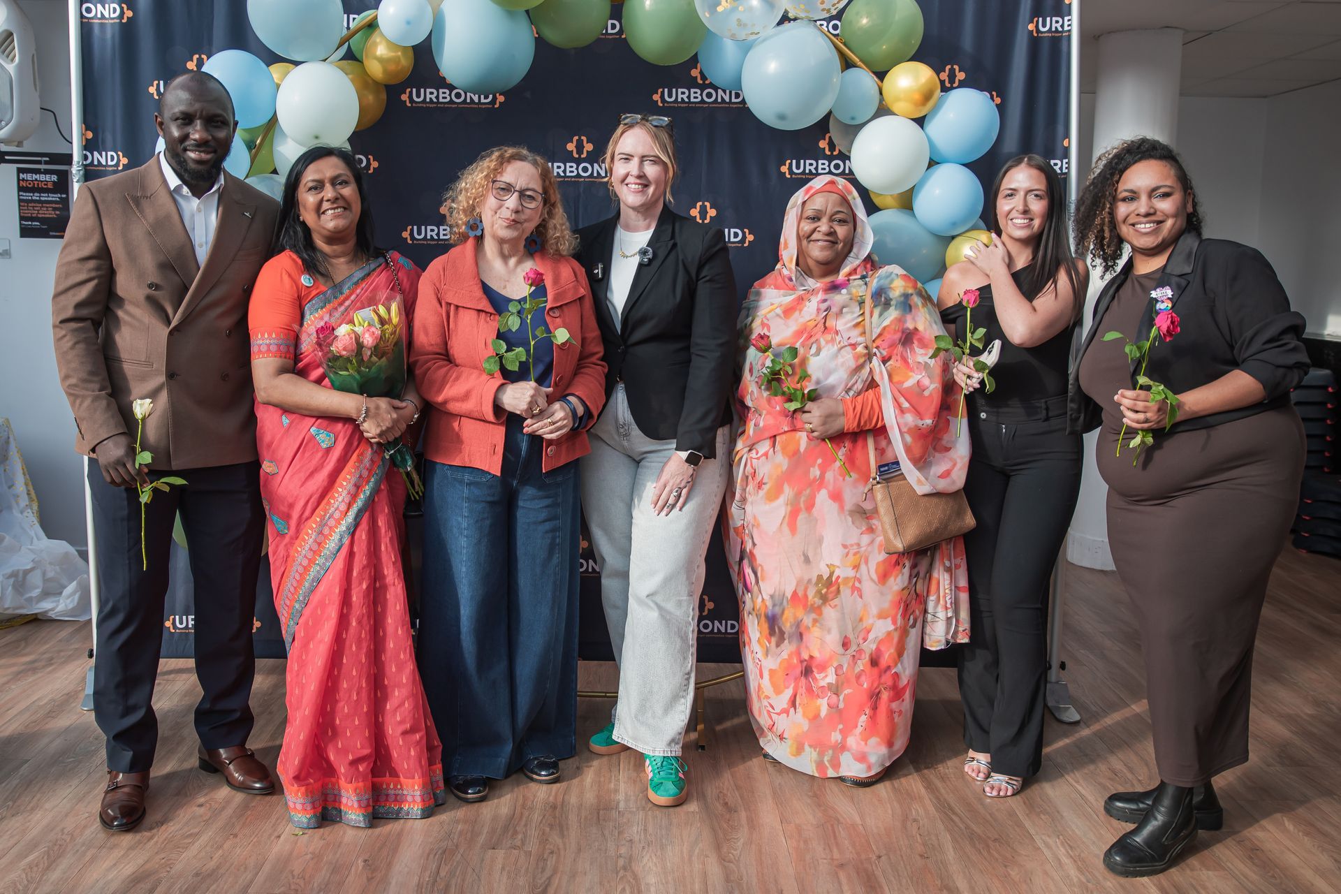 Seven people stand in a line posing for a photo against a backdrop of balloons, smiling and holding small bouquets.