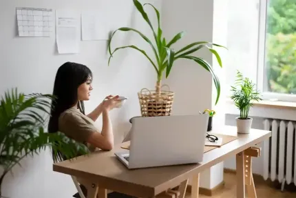 Woman at desk with laptop, gesturing with phone, plants in bright room.