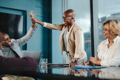 People in a business meeting giving high five. Woman in a tan blazer, man in a grey suit, and another woman smiling.