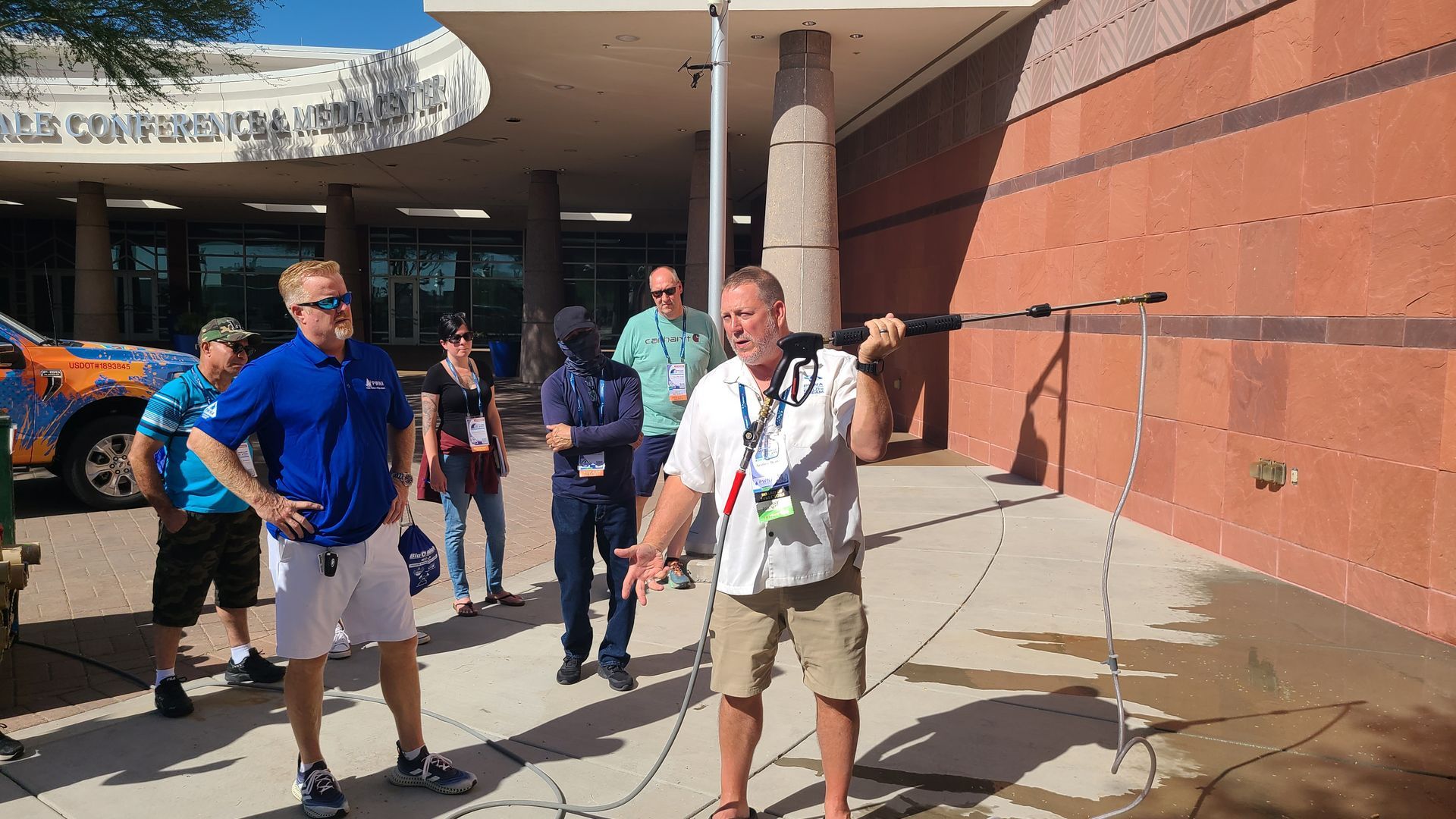 People standing outside a building entrance, including one man speaking into a microphone on a sunny day.
