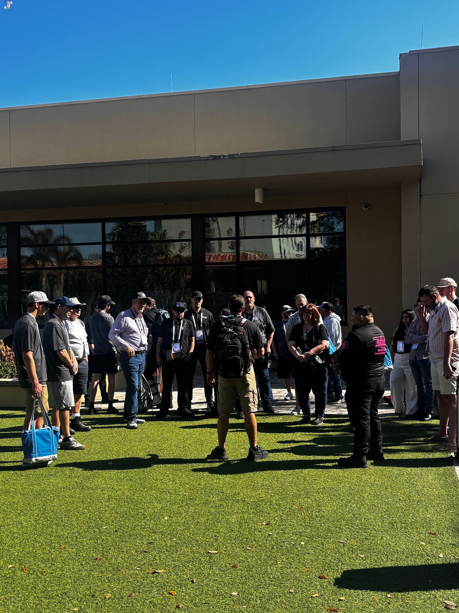 Group of people gathered outside a building on a sunny lawn, some holding bags and talking.