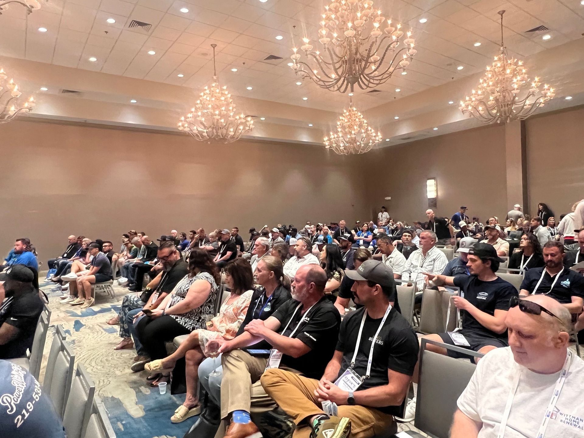 Audience seated in a large conference ballroom with chandeliers and rows of tables and chairs