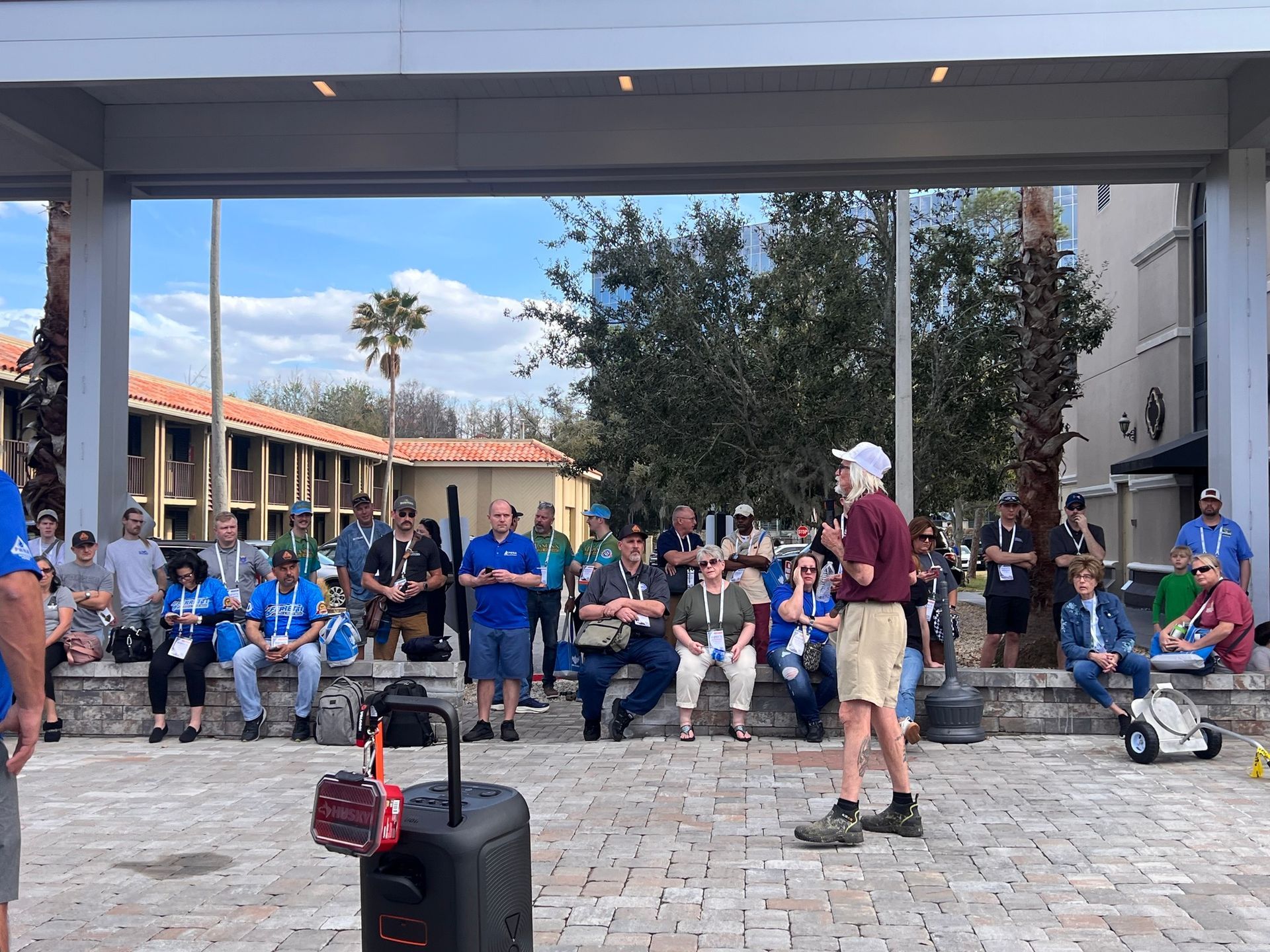 Crowd seated and standing in a covered courtyard, with luggage and a man in maroon shirt in the foreground