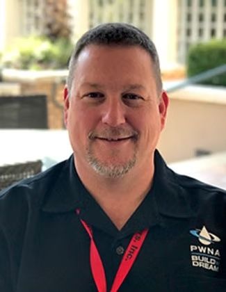 Smiling man in black polo and red lanyard standing outdoors with blurred buildings in background