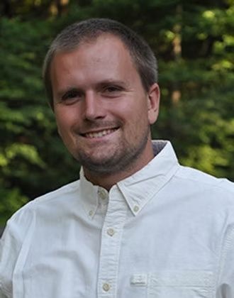 Smiling person in a white button-down shirt outdoors with blurred green trees in the background