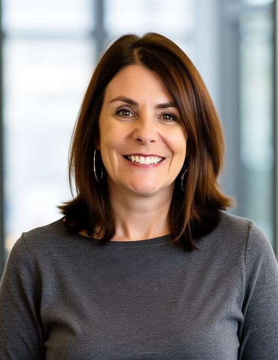 Smiling woman in a gray top standing indoors against a bright windowed background
