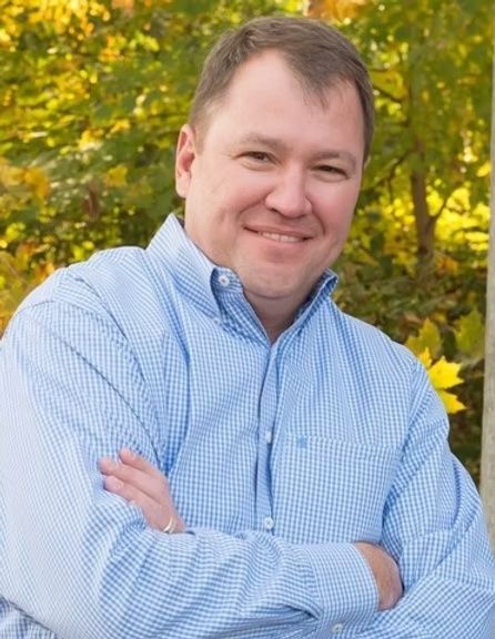 Smiling person in a blue button-down shirt with arms crossed, standing outdoors with autumn foliage.
