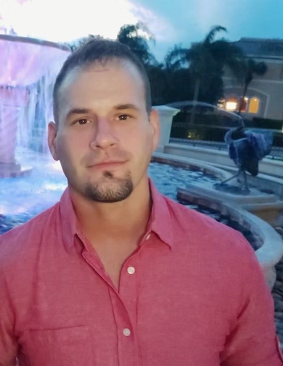 Man in a pink shirt standing by a lit fountain at dusk, with palm trees in the background