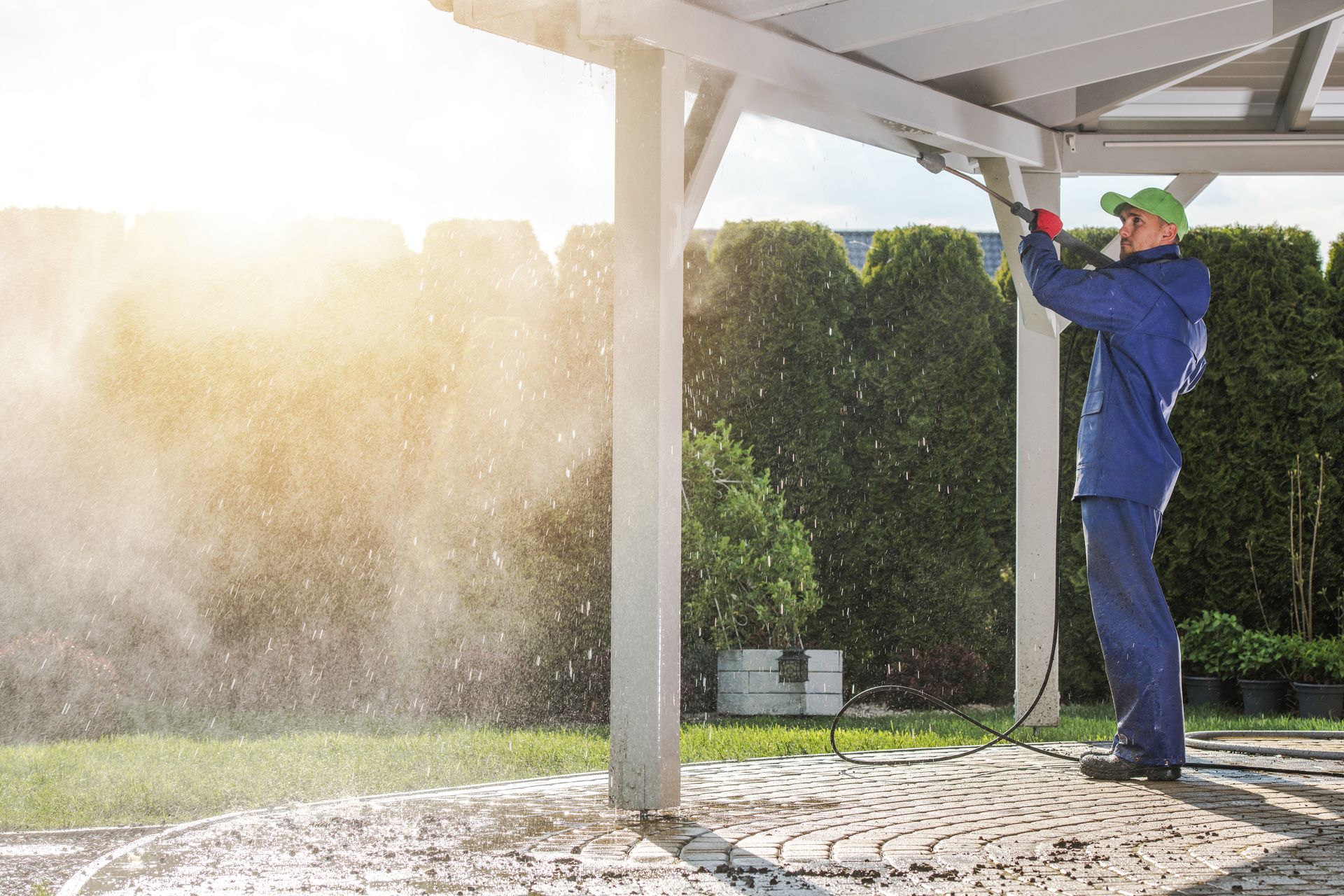 Person pressure-washing a patio beside a hedge, spraying water under a pergola in bright sunlight