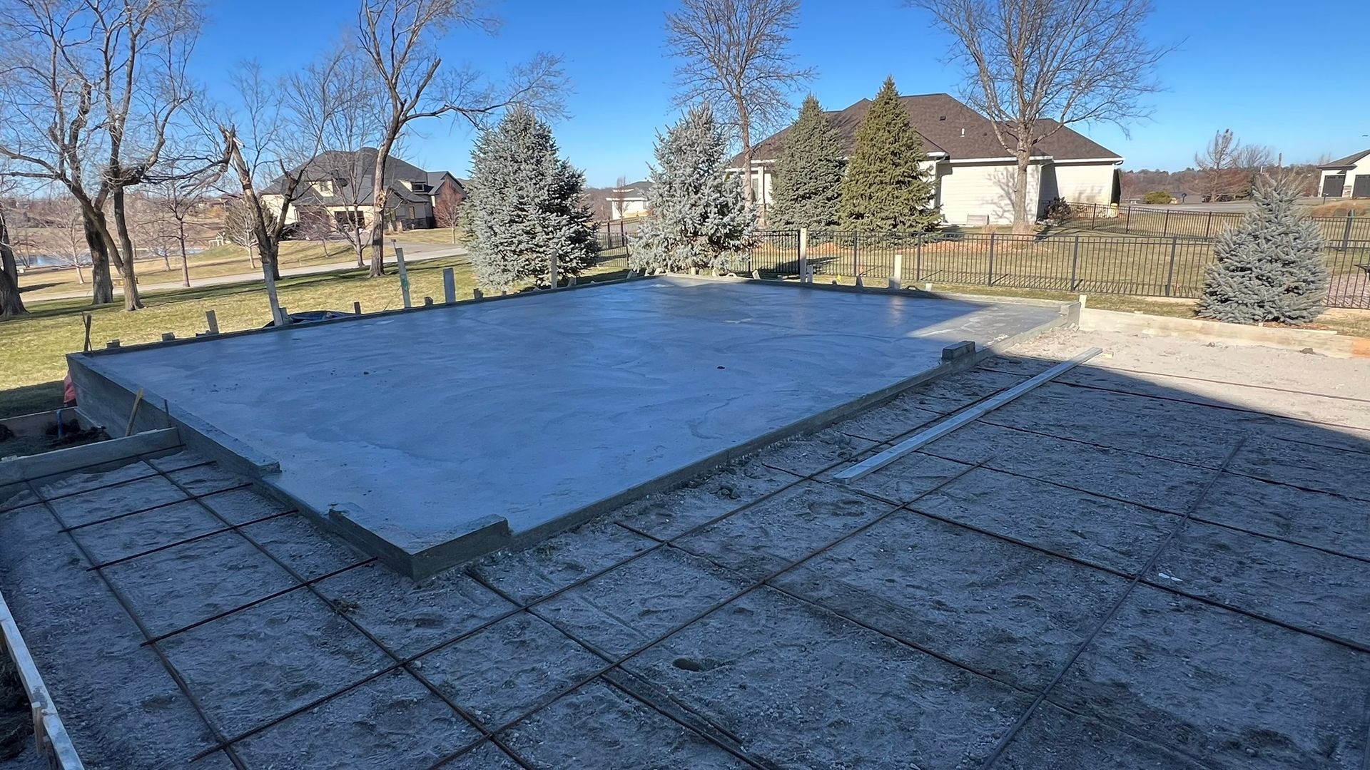 Construction site with concrete foundation, soil, and houses in the background on a cloudy day.