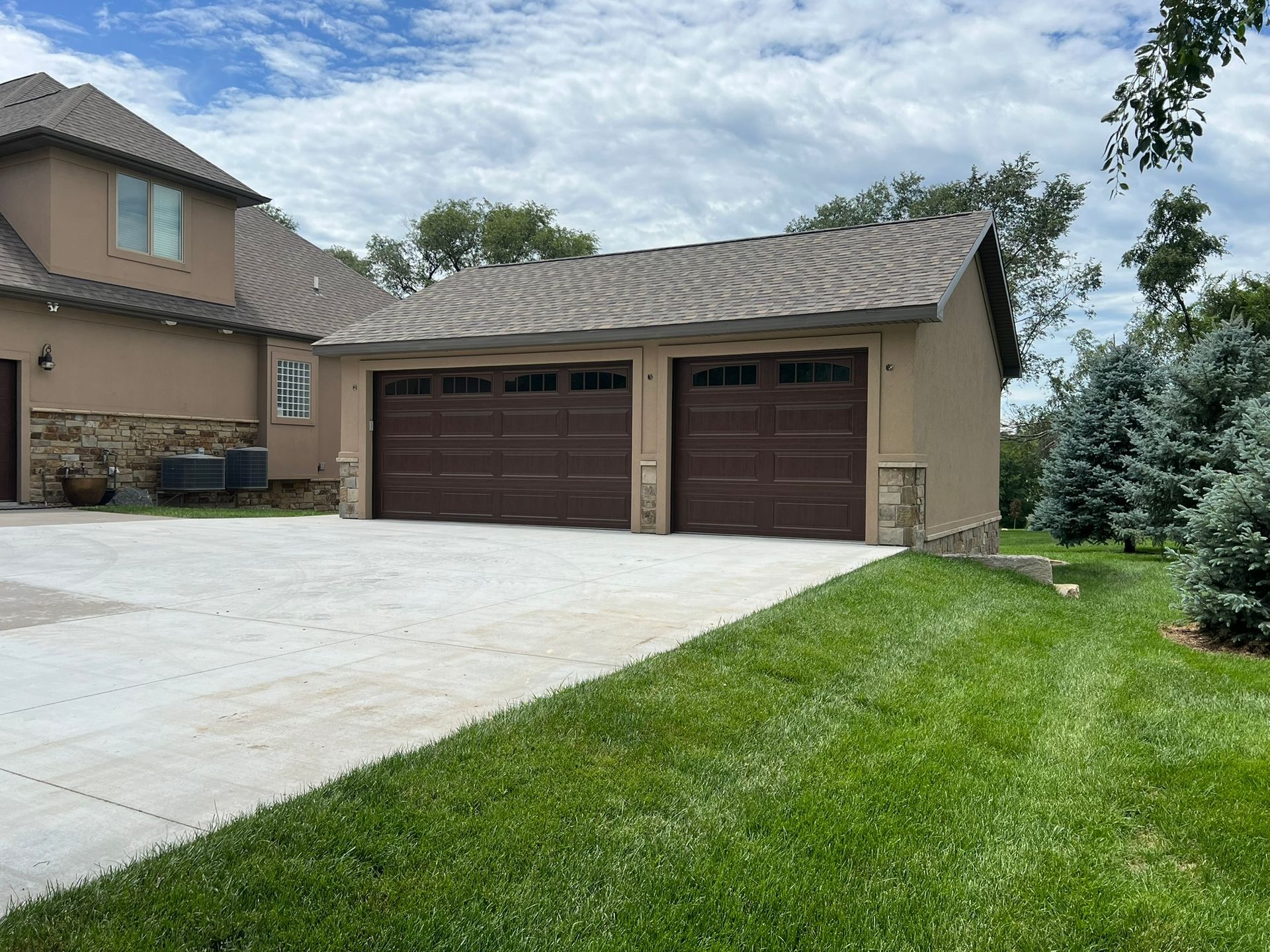 Two-car garage with brown doors, tan stucco exterior, set next to a house, with grass and driveway.