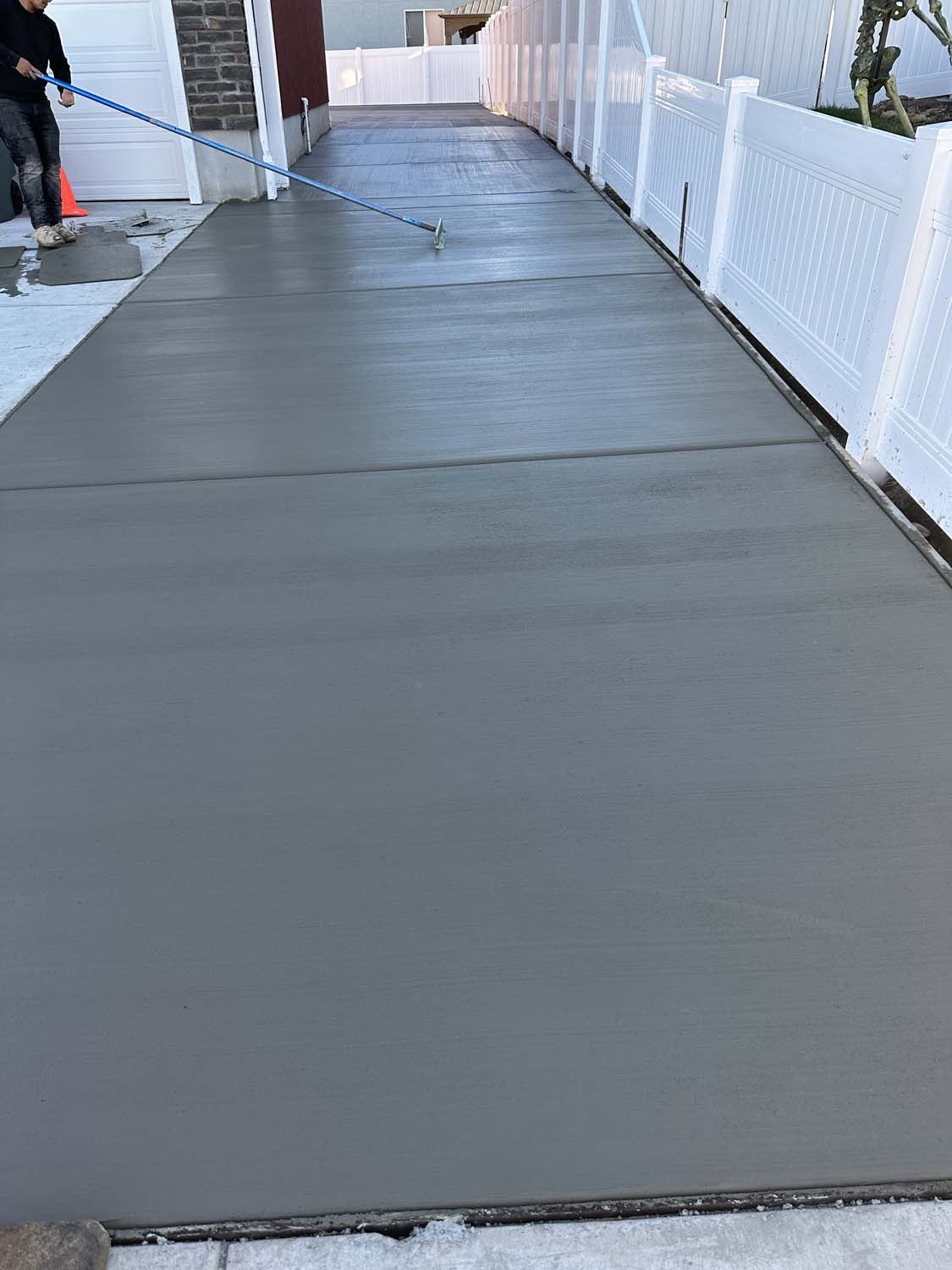A man is laying concrete on a driveway next to a white fence.