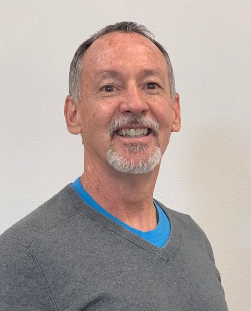 A man with a beard is wearing a grey sweater and a blue shirt.