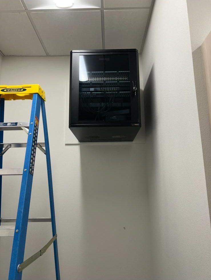 Blue ladder in a hallway beside a wall-mounted black electrical panel under ceiling tiles.
