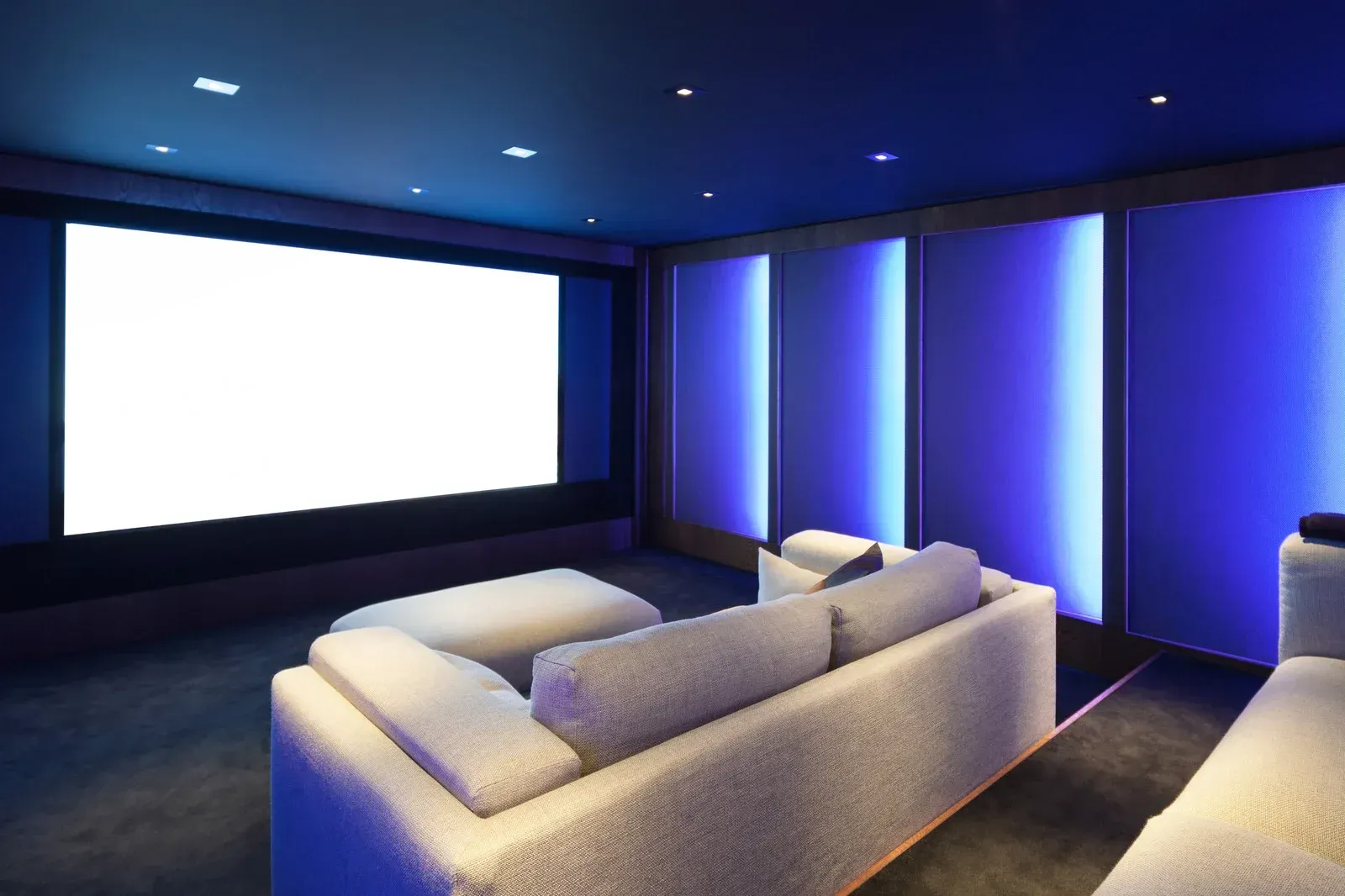 Modern home theater with a large white screen, beige sectional sofa, and blue LED accent lighting