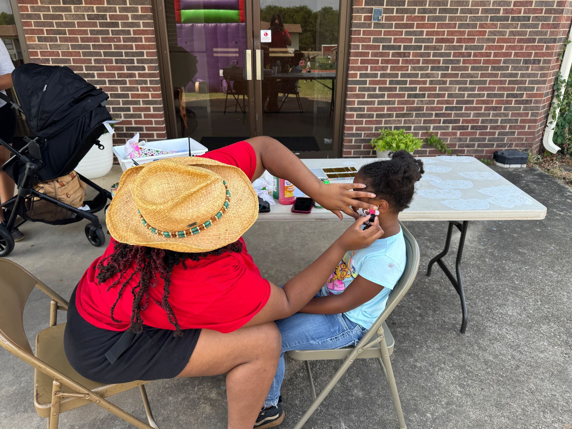 Person painting a child's face outdoors; the child sits on a chair in front of a brick building.