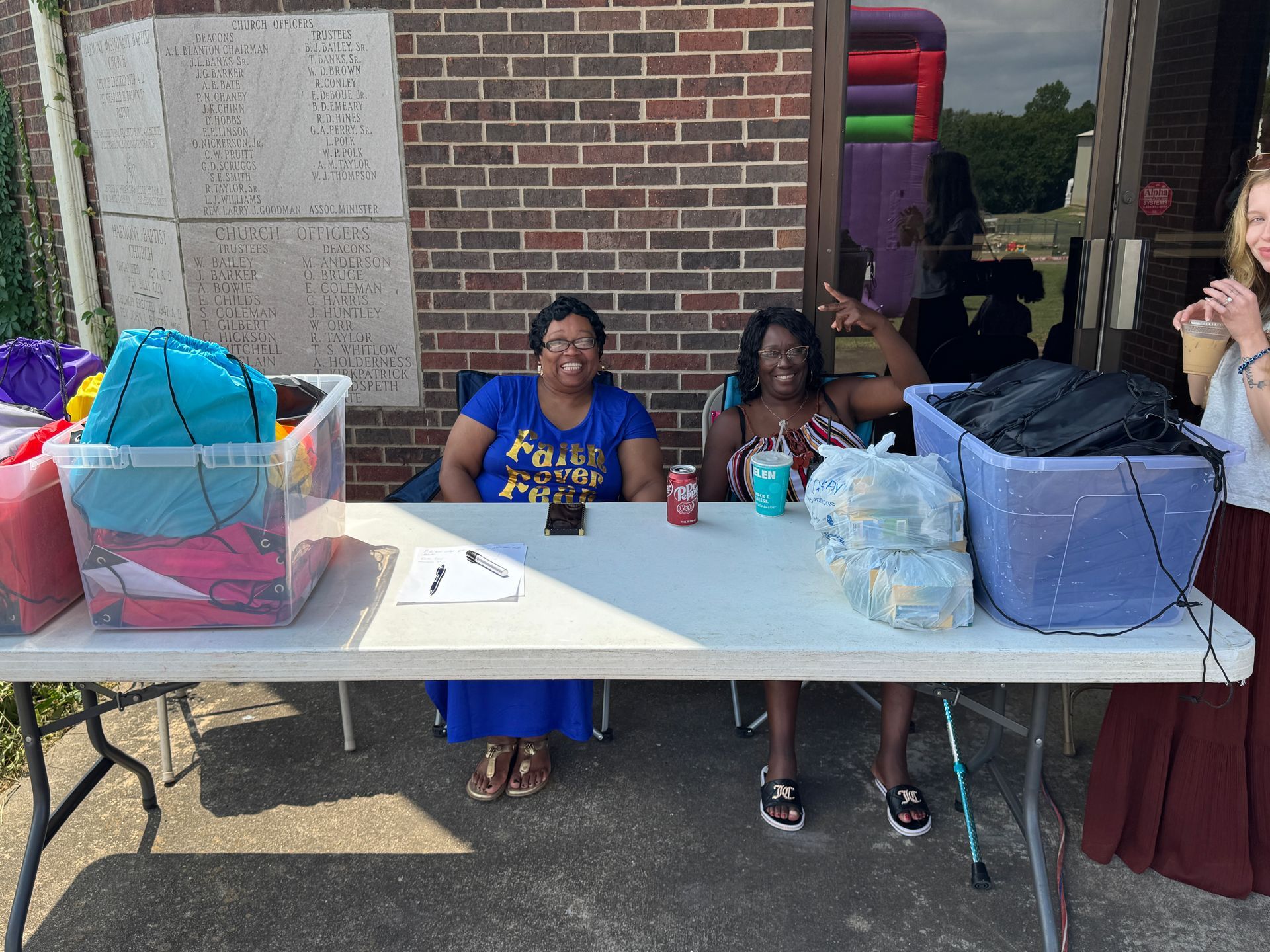 Two women seated at a table, smiling, near a brick building with a bouncy house in the background.
