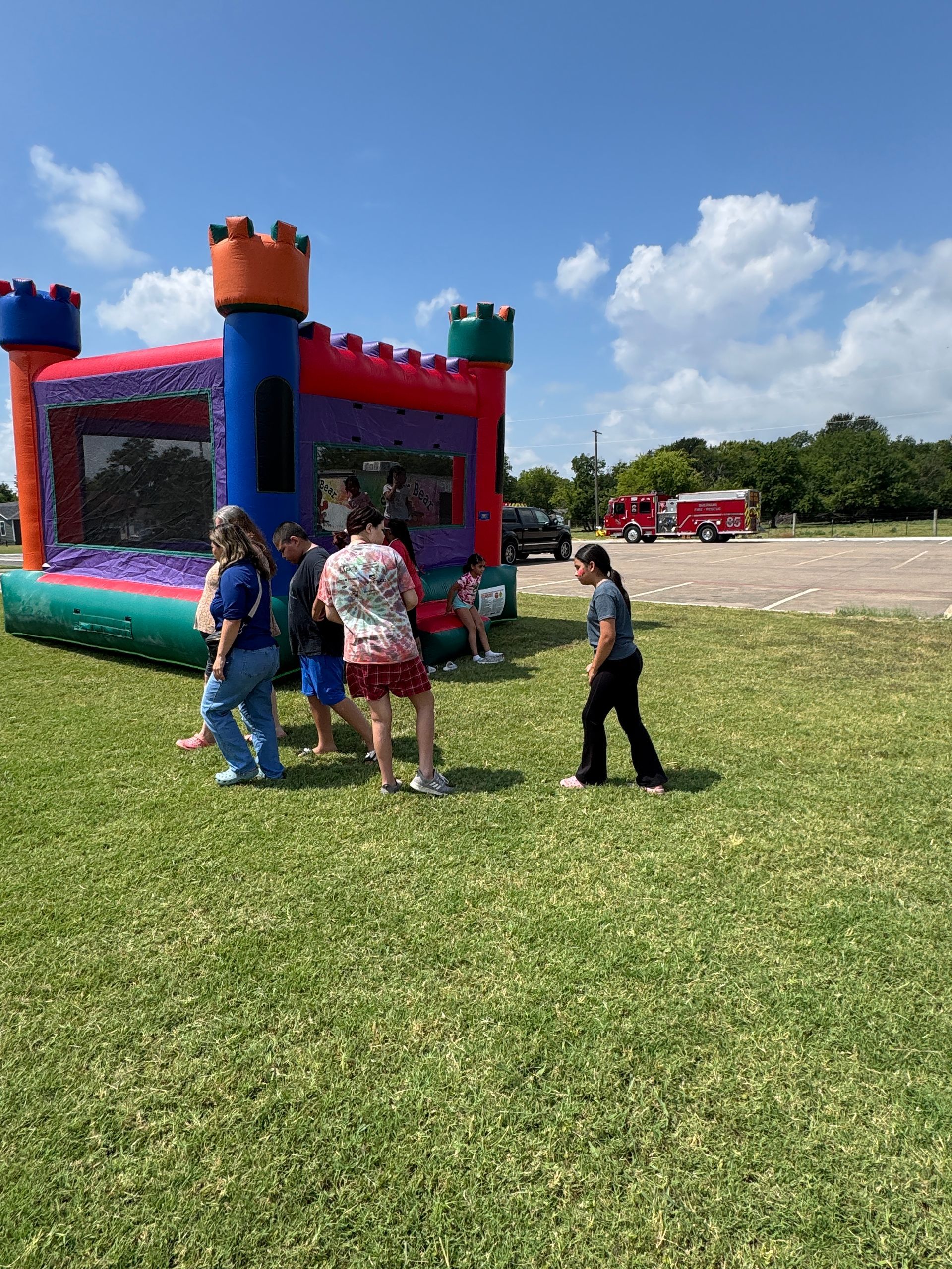 Kids and adults near a colorful castle-shaped bouncy house on a grassy area under a blue sky.