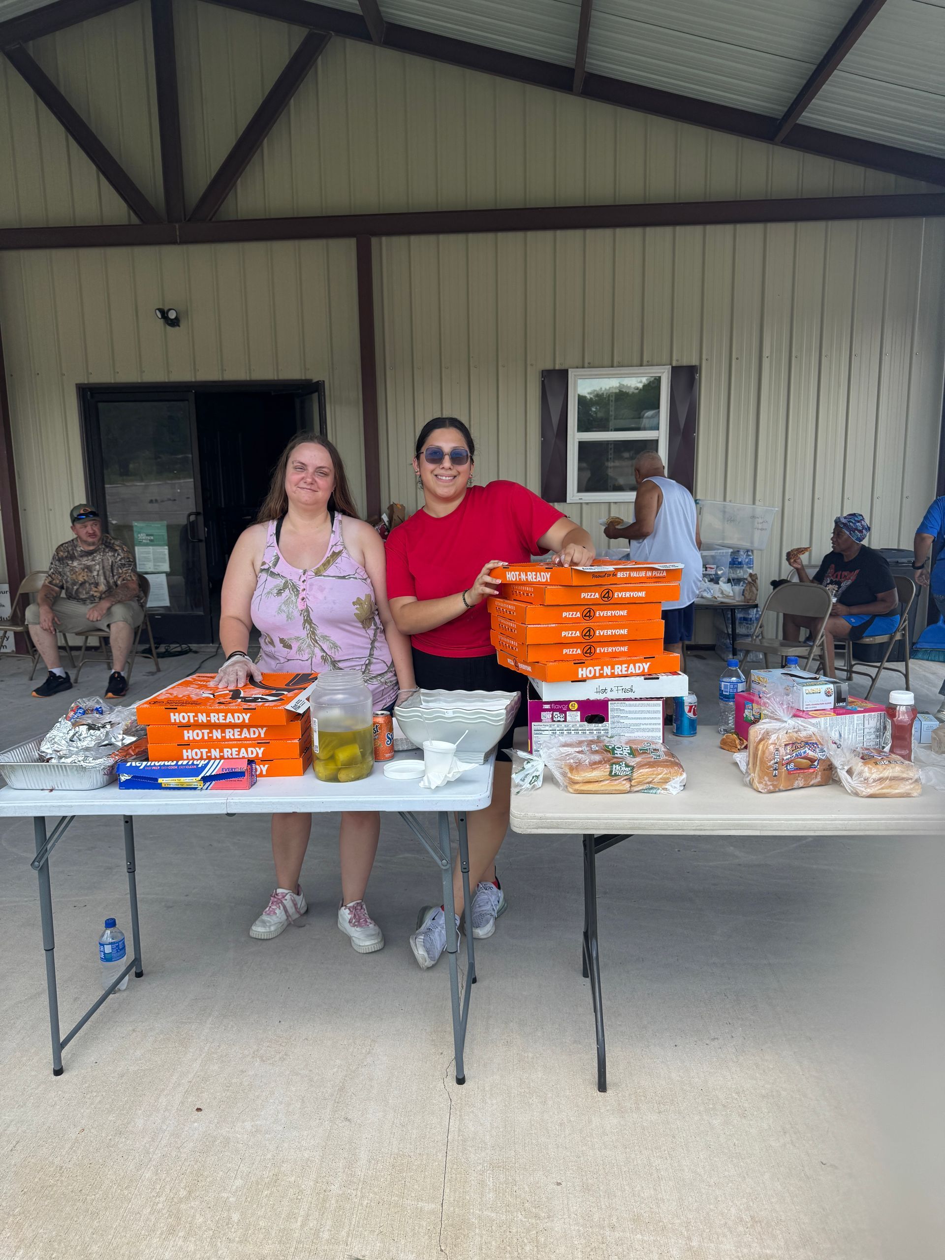 Two women at a picnic table with food, one in a red shirt, outside a building.