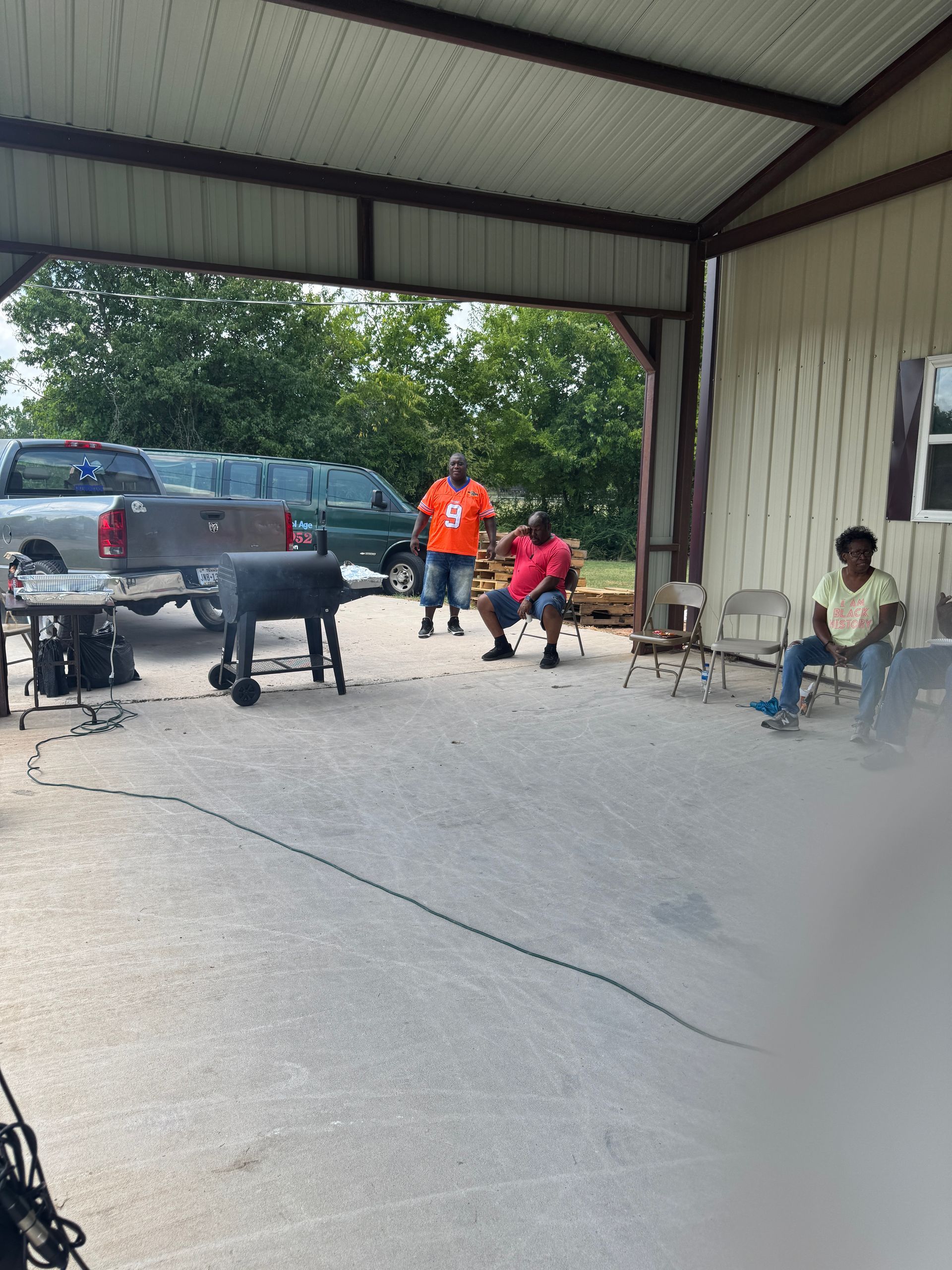 People gathered under a carport; one grilling, others sitting. Trucks and trees in the background.