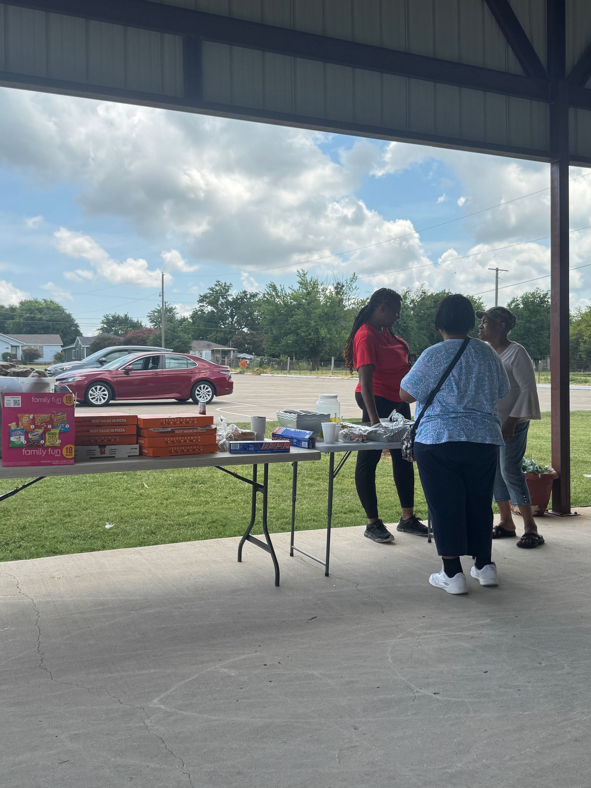 People at a table under a pavilion. A woman in red serves others. Red car and trees in the background.