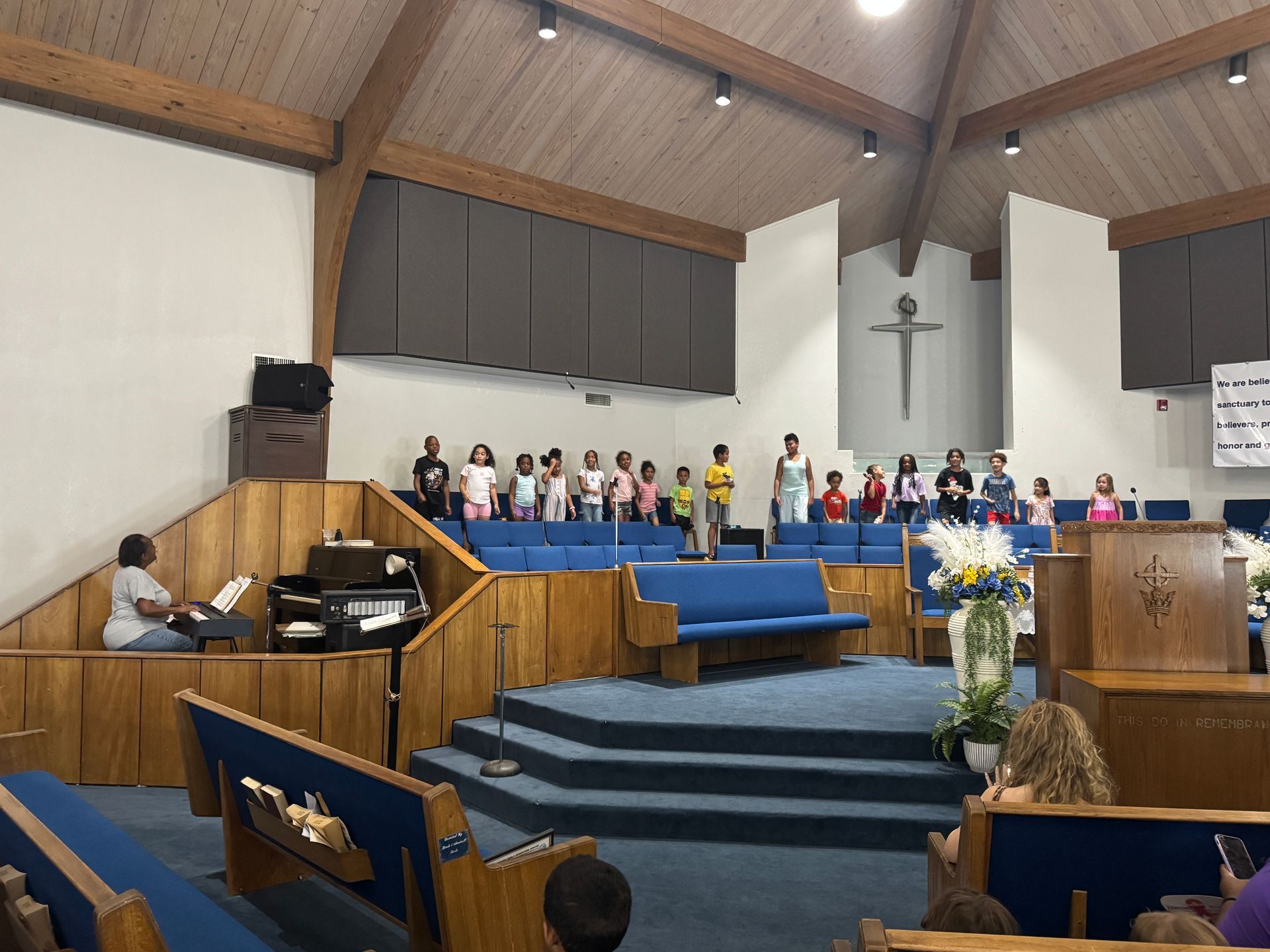 Children's choir performs on a stage inside a church. Blue seating, wooden accents, cross on the wall.