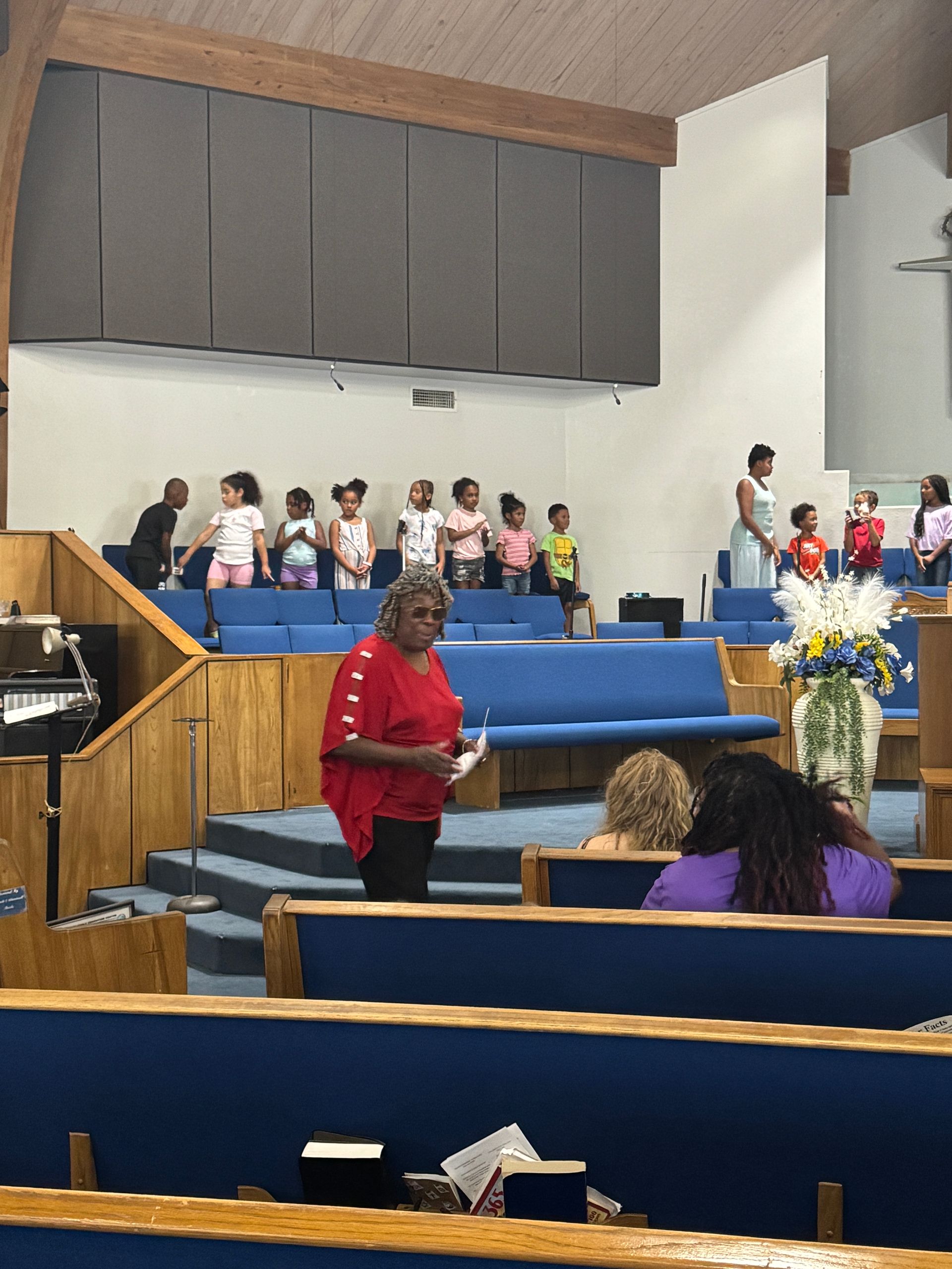 Woman in red top speaks to children on stage in a church.