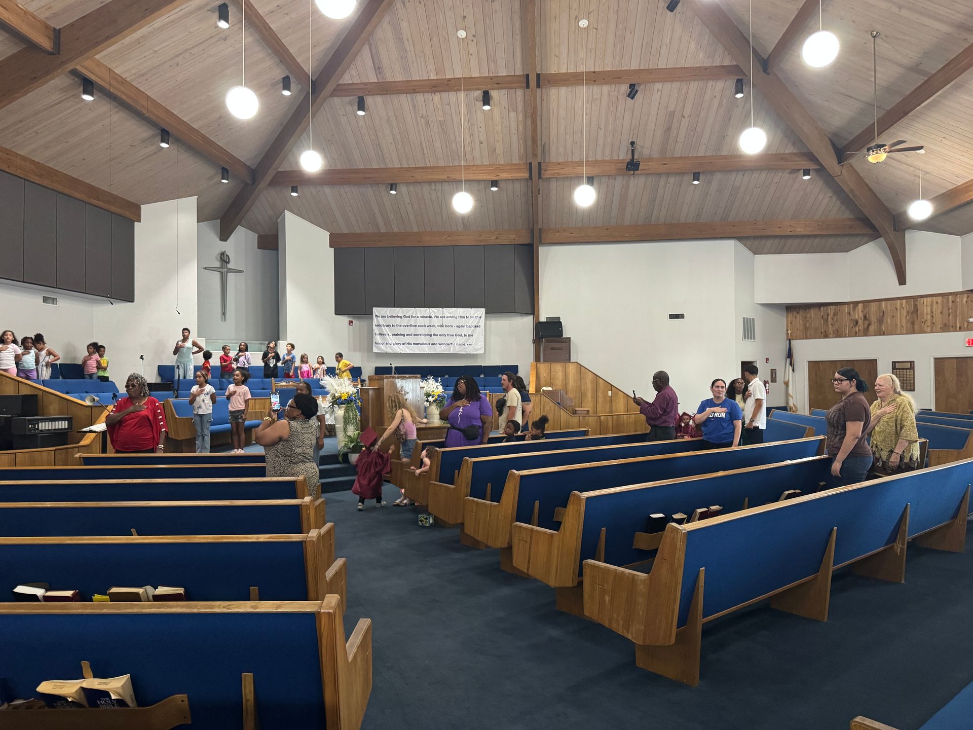 Interior view of a church service; people standing near pews. Wooden beams and a cross are visible.