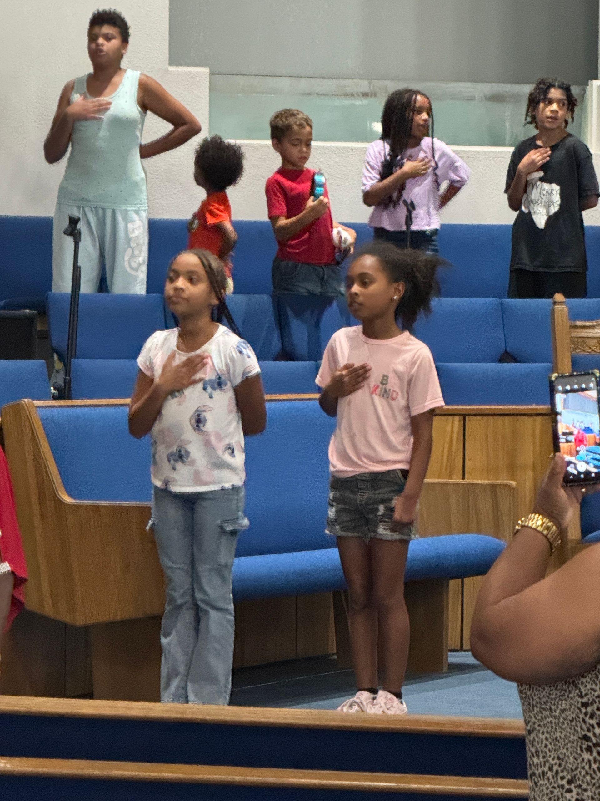 Children and adults stand with hands over hearts in a church, reciting the Pledge of Allegiance.