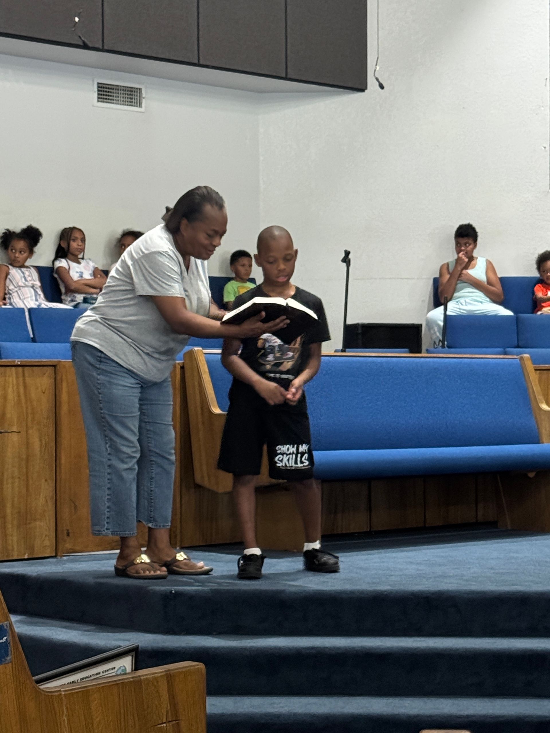 Woman helps boy read a book on a church stage; others sit in blue pews.