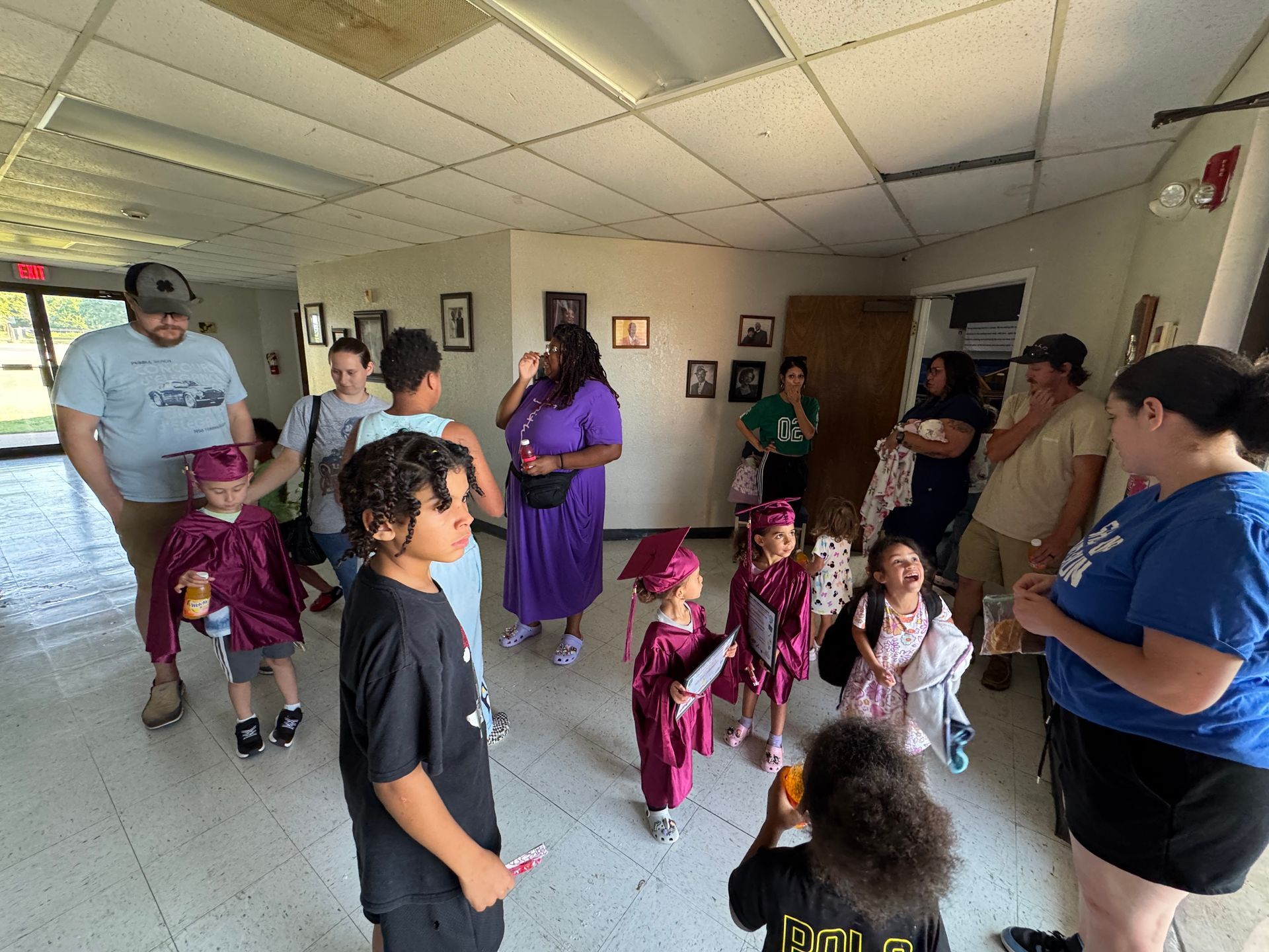 Children in graduation gowns with adults in a bright room, likely a school or event.