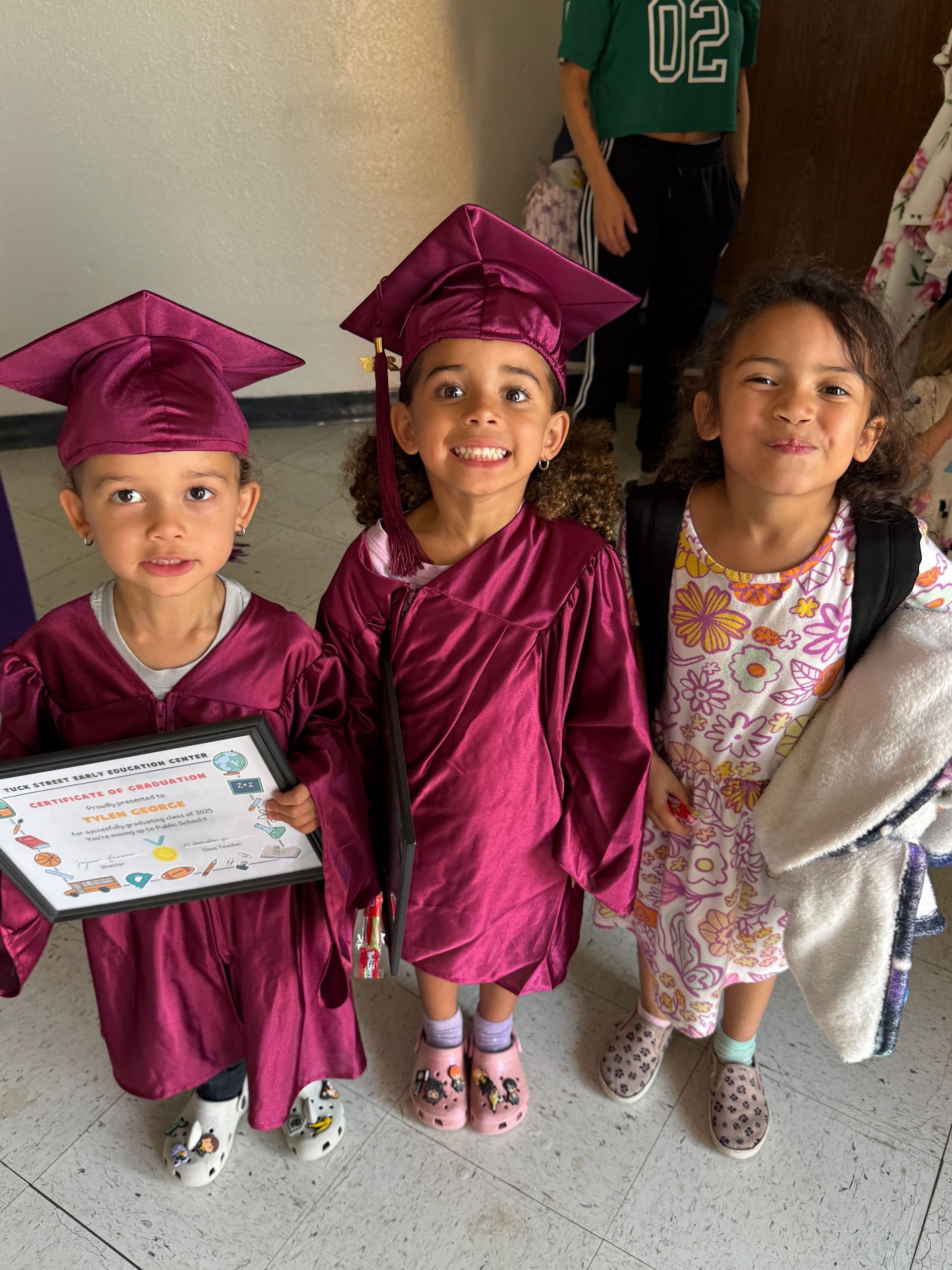 Three young children in maroon graduation gowns smile. One holds a diploma.