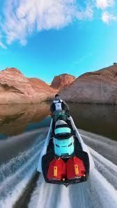 A person rides a jet ski across calm water toward red rock formations under a bright blue sky.