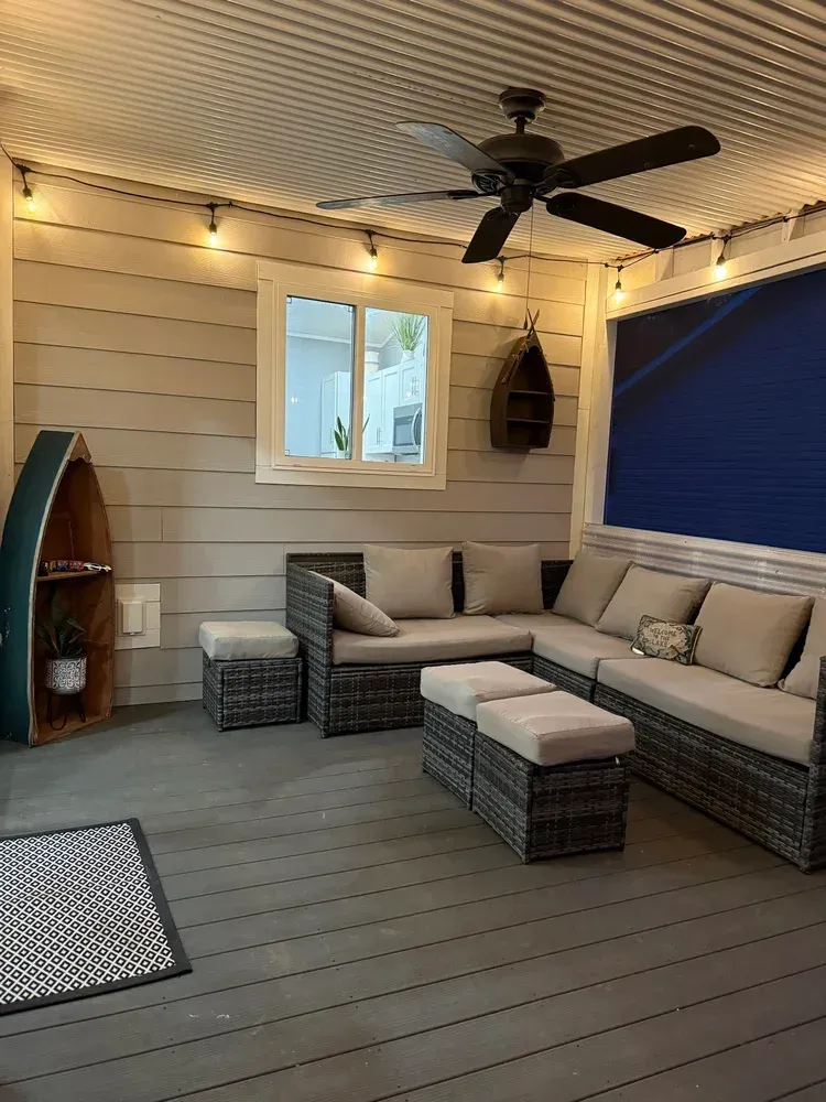Covered patio with beige sectional, wicker ottomans, string lights, ceiling fan, and blue privacy blind