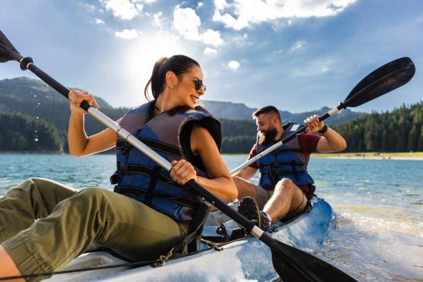 Two people kayaking on a lake, paddling with mountains and trees in the background.