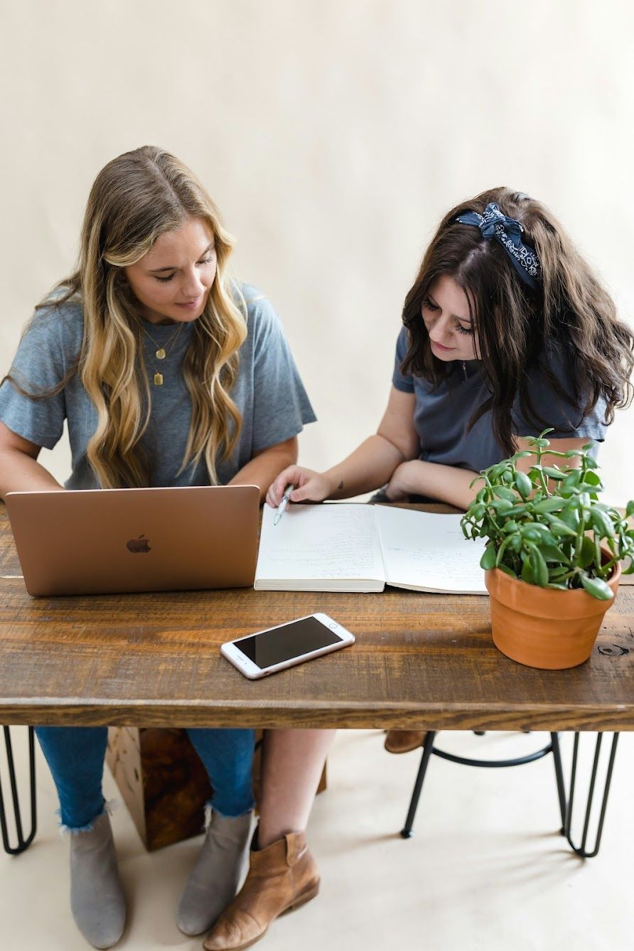 Two girls are sitting at a table with a laptop and a book.